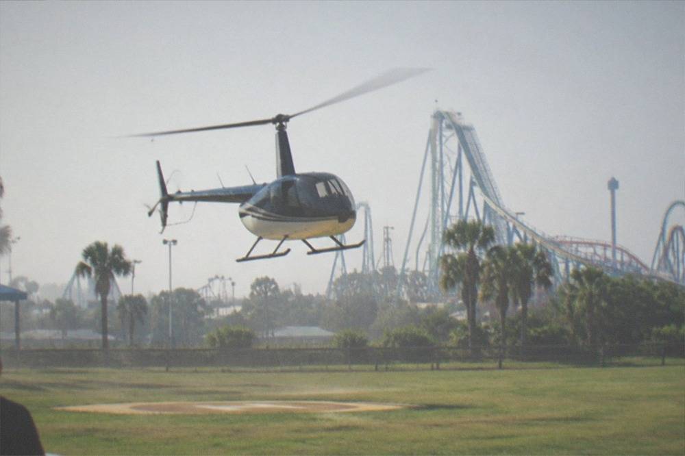 A helicopter lifting off from a grassy helipad with theme park area in the distance