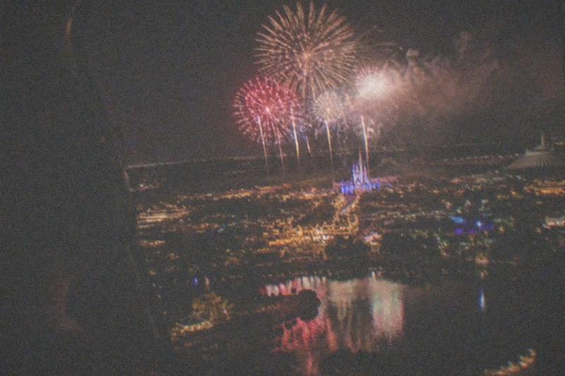 Colorful fireworks blooming above distant theme park rooftops, watched from a helicopter cabin after dark, the bursts mirrored in lakes far beneath, no people, no logos, no readable text.