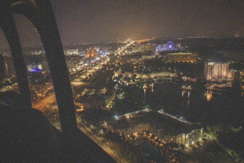 Nighttime helicopter-cabin view over a brightly lit Orlando boulevard, streams of headlights and glowing hotel signs set against dark central Florida flatland, no people, no logos, no readable text.