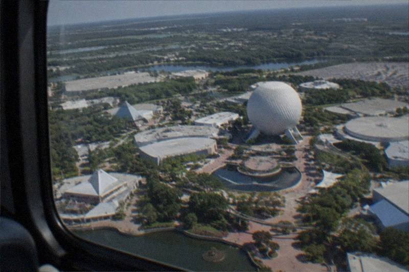 Daytime helicopter-cabin perspective above the Orlando park corridor, colorful rooftops of distant attractions and blue lakes stretching toward the horizon under sunshine, no people, no logos, no readable text.