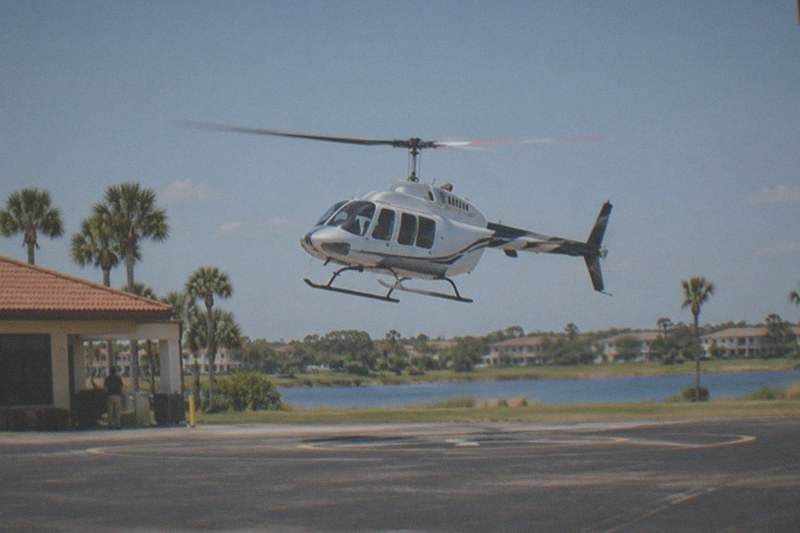 Tour helicopter lifting off from a Kissimmee-area pad into clear afternoon sky, flat suburban landscape with scattered lakes passing below the rotor, no people, no logos, no readable text.
