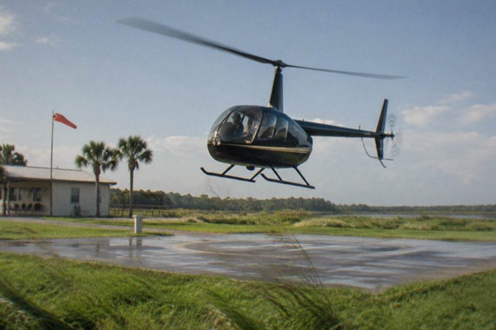 A small tour helicopter lifting off from a grass-edged helipad in flat morning light