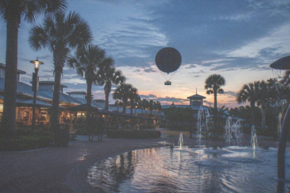 A waterfront promenade with hot air balloon silhouette, shops and palm trees at dusk