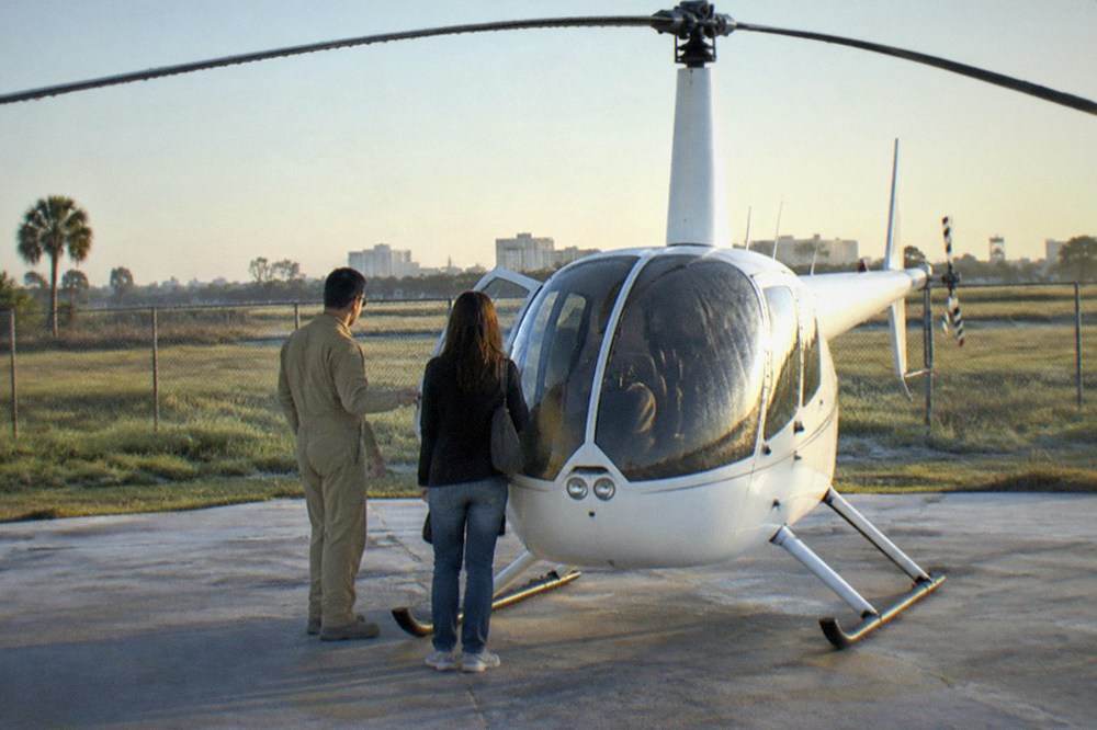 Pilot and passenger in flight suits standing beside a helicopter on a clear morning at a Kissimmee helipad