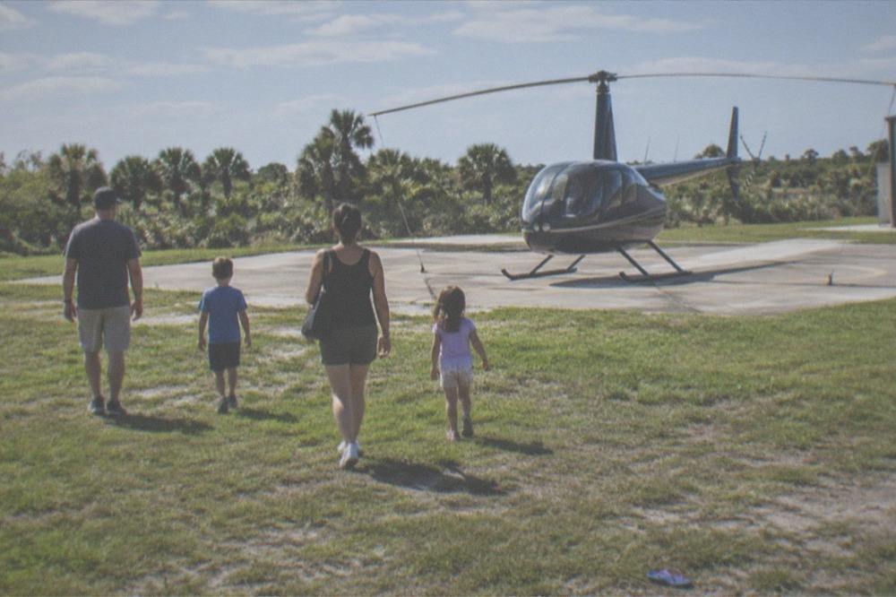 Family walking toward a small helicopter across a sunlit helipad lawn