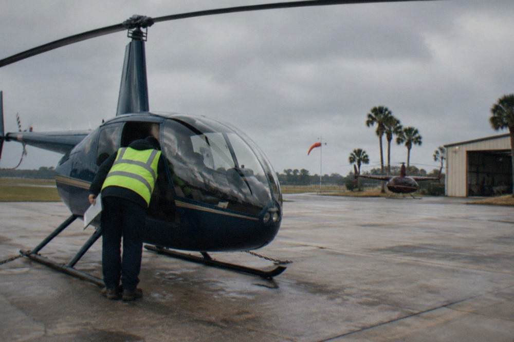A small sightseeing helicopter parked on a helipad with a crew member completing a pre-flight checklist.