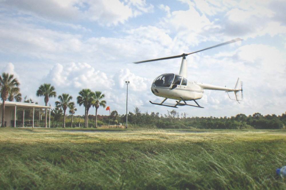 A small helicopter lifting off from a grassy pad under a partly cloudy sky