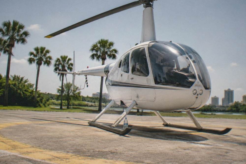 A small sightseeing helicopter parked on a paved helipad with palm trees and open sky behind it