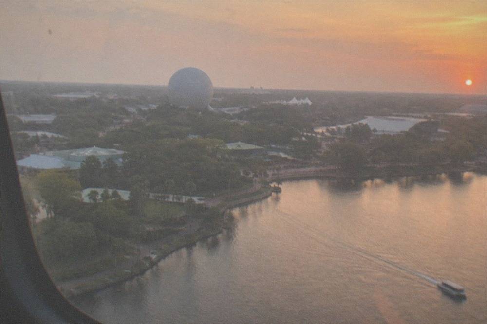 Aerial view from a helicopter window showing a lake shoreline and distant theme park structures