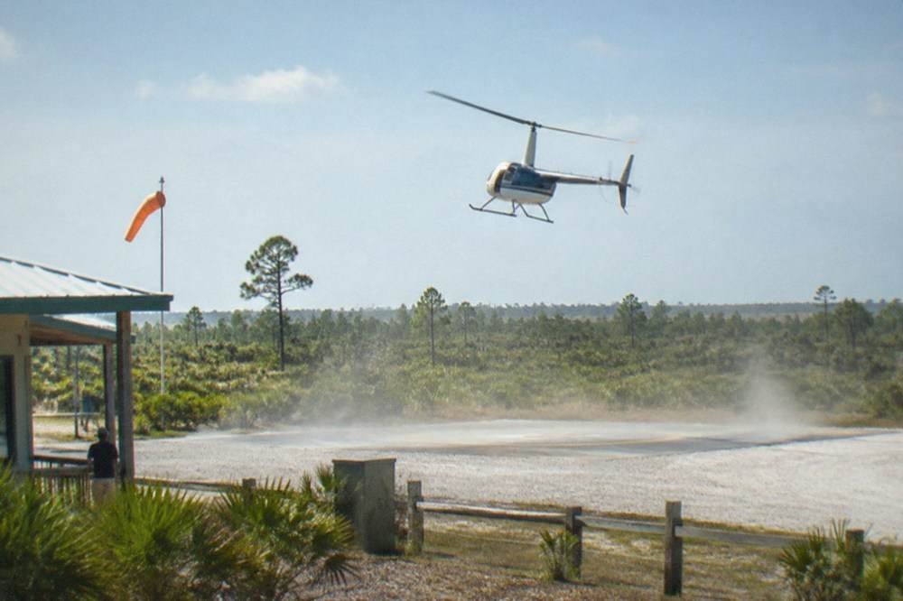 A light helicopter climbing away from a small Kissimmee heliport with flat Florida terrain stretching to the horizon