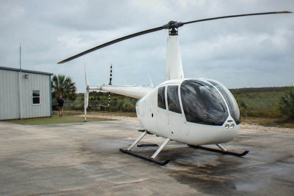 A light tour helicopter parked on a paved helipad next to a small Florida airport building