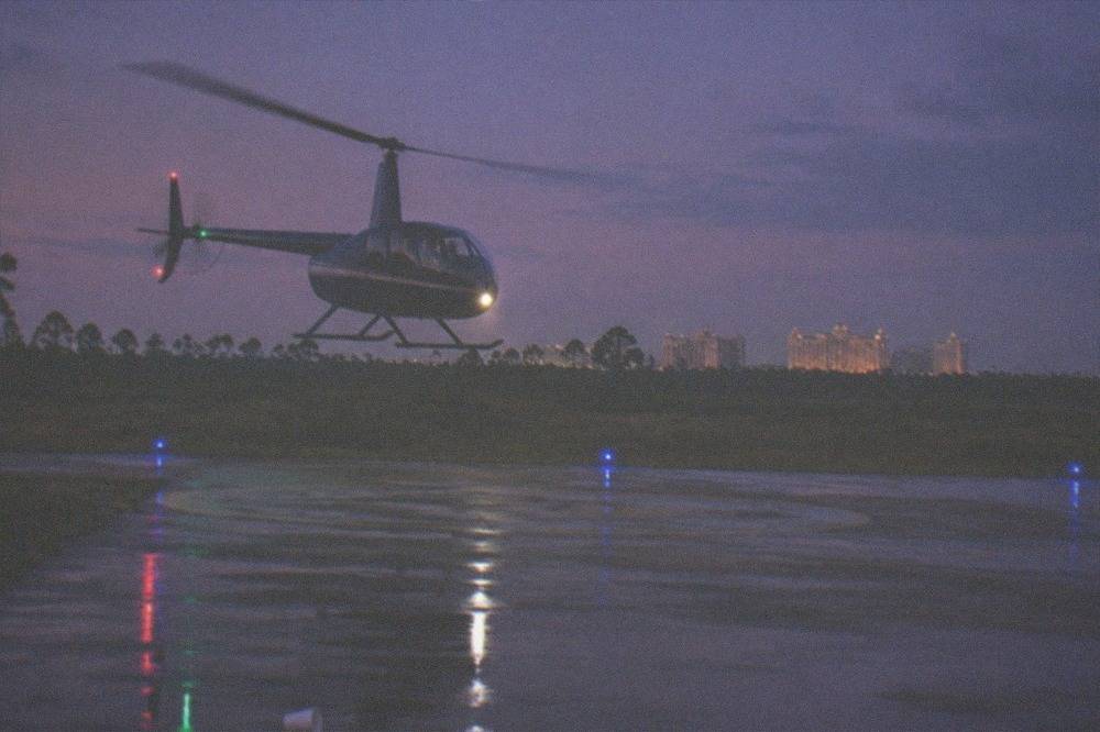 A tour helicopter lifting off a helipad with a resort skyline and dusk sky in the background