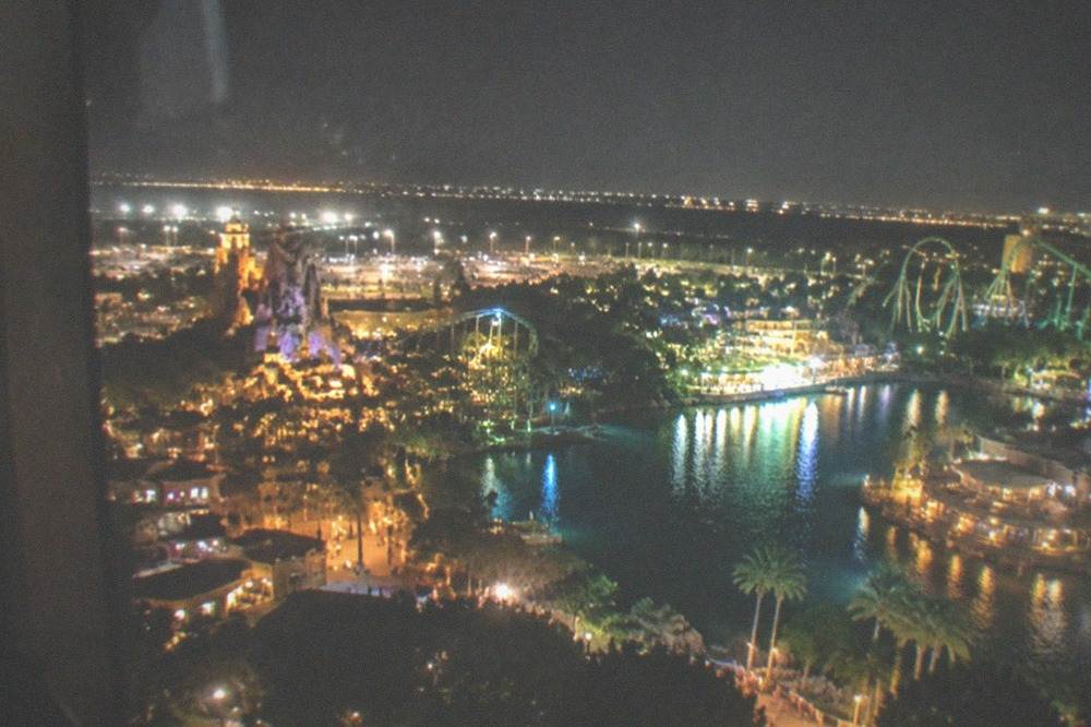 Lit theme park area at night seen from a helicopter with city lights spread below