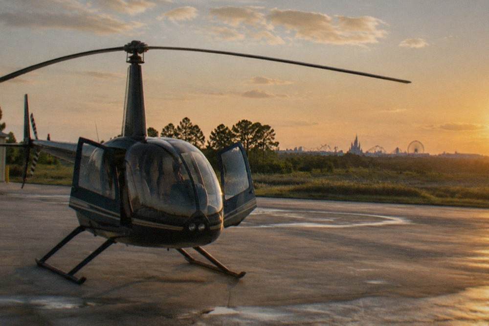 A four-seat Robinson helicopter on a paved apron with theme-park skyline in the far distance