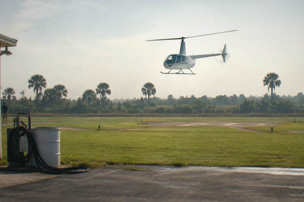 A helicopter lifting off from a grassy Kissimmee helipad with palm trees along the horizon