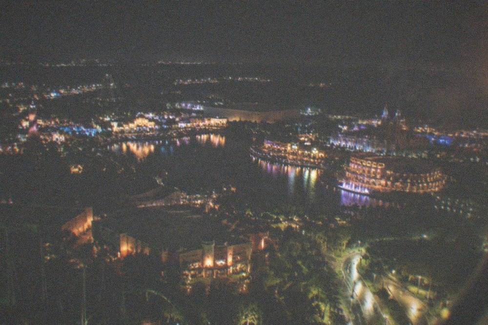 Night aerial view of a brightly lit city skyline with illuminated roads and distant fireworks glow