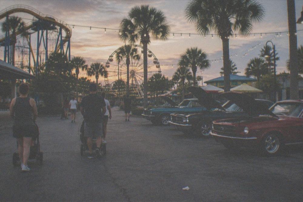 Outdoor evening promenade with string lights, palm trees and people strolling past restaurants