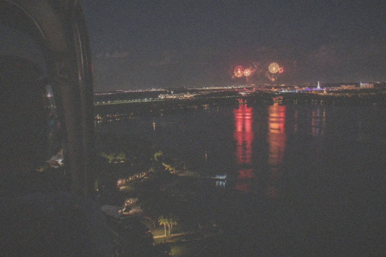 Aerial night scene above the Orlando resort corridor. From a helicopter window, bright firework shells bloom in red and gold over distant park skylines while their reflections scatter across the lakes of Kissimmee and Lake Buena Vista, empty cabin, no branding, no lettering.