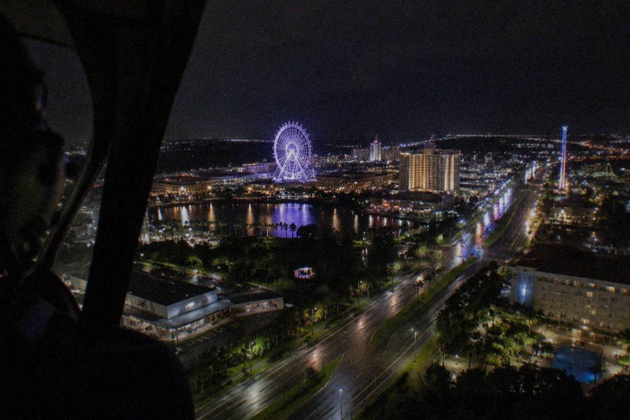 Wide establishing view. Night helicopter-cabin panorama above the International Drive corridor, a wide glowing band of roadway, hotels, and signage beneath a black sky, no people, no logos, no readable text.