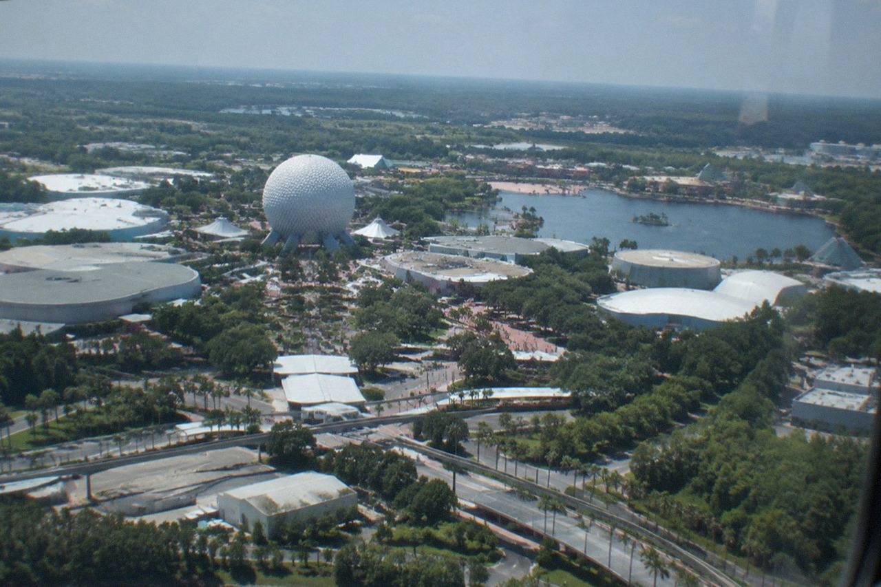 Wide establishing view. Daytime aerial view from a helicopter cabin over the Orlando theme-park corridor, distant colorful rooftops and lakes stretching to a bright horizon, no people, no logos, no readable text.